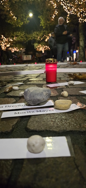 Nahaufnahme von Kopfsteinpflaster bei Nacht,
 auf dem kleine Steine als Beschwerer auf weißen Papierstreifen mit Namen wie Moritz Mayer liegen. Im Hintergrund leuchtet eine rote Grabkerze,
 während im unscharfen Hintergrund Menschen unter beleuchteten Bäumen stehen.